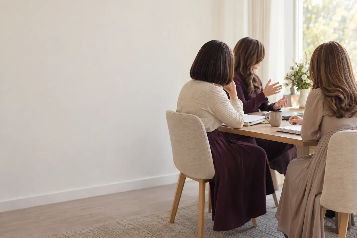 Three women learning together around a table with notebooks, in bright window light